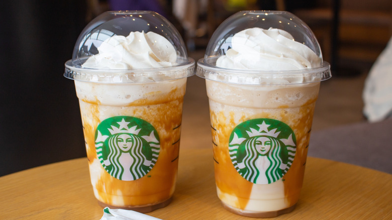 Two caramel frappuccinos with whipped cream and drizzle around the cups sit on a table