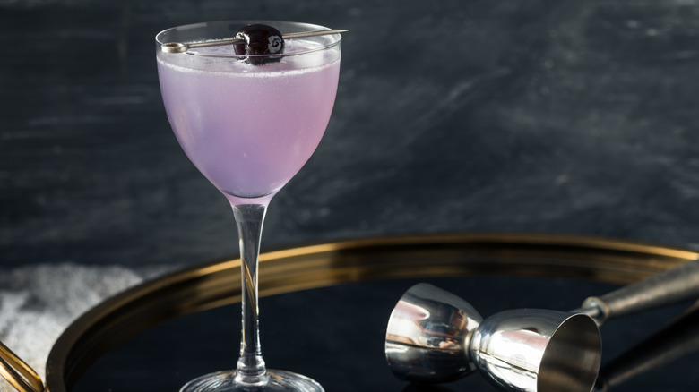 A purple aviation cocktail with a clack olive, on a tray, with a measuring jigger