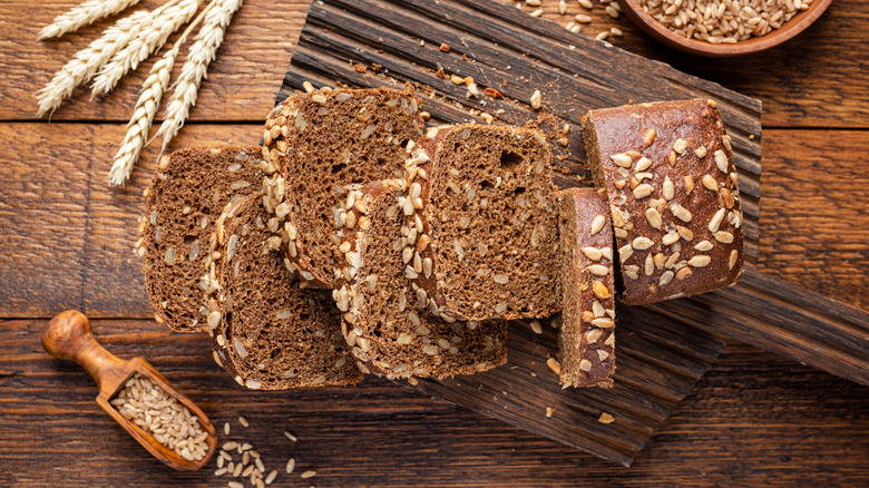 Slices of dark bread containing seeds and whole grains sit on a wooden baker's peel, with containers and stalks of grains in the background
