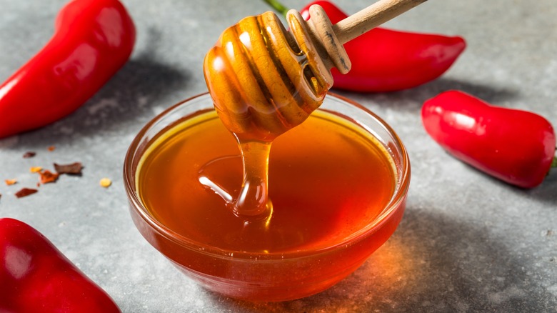 Honey dipper dripping reddish honey into a small glass bowl, surrounded by hot red peppers on a gray surface
