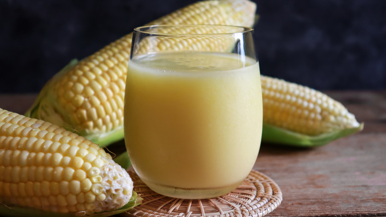 Corn milk is a clear glass, sitting on a wicker coaster with ears of corn on the table surrounding it