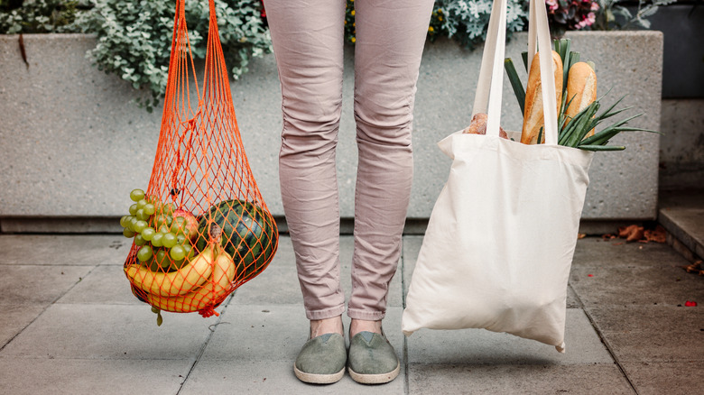 person holds two reusable grocery bags with food while walking