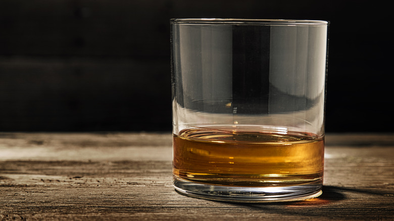 a glass with a small amount of bourbon sits on wooden surface