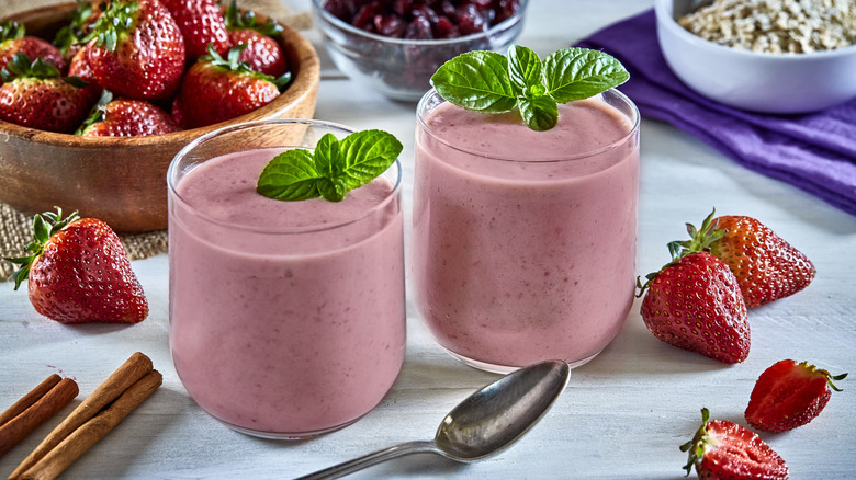 Two pink smoothies in a glass on a table, garnished with mint sprigs, surrounded by a bowl of strawberries, oatmeal, cinnamon sticks, individual strawberries, and a spoon