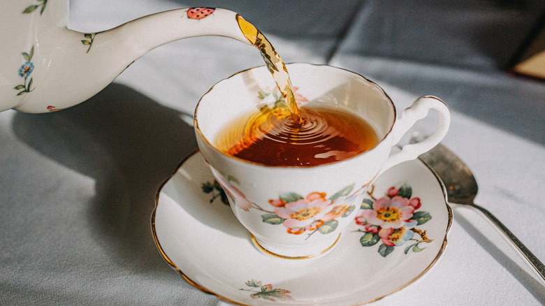 Teapot pouring black tea into a china teacup on a small serving plate