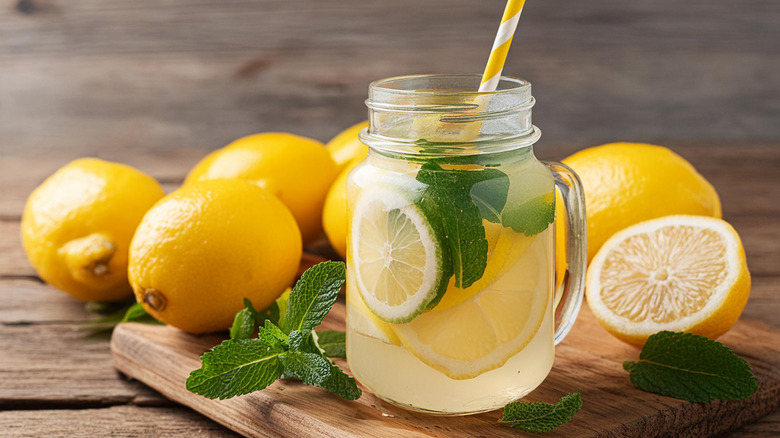 A cup of lemonade with lemon slices and mint on a wooden cutting board with whole lemons and mint leaves.