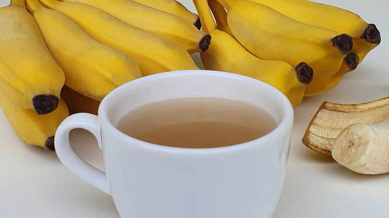 White tea cup with tea inside, and two bunches of bananas in the background. Single peeled banana to the right side of tea cup.