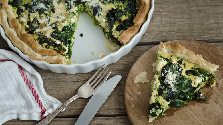 A cheese and spinach quiche in a pie pan, with one slice removed next to a fork and knife