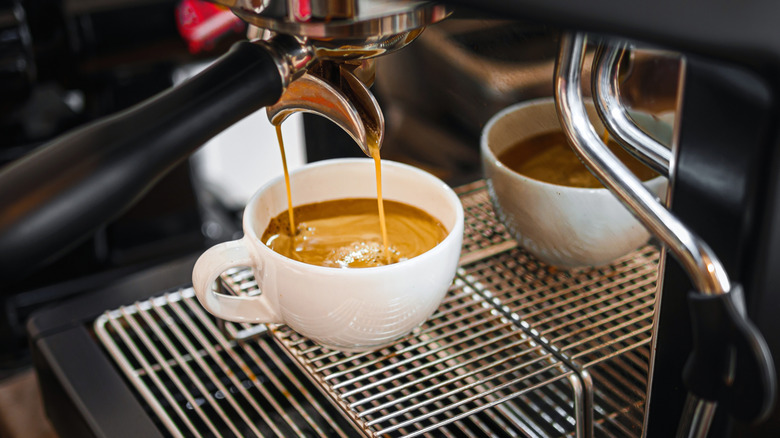 A white mug of espresso on an espresso machine, with coffee being poured into the mug