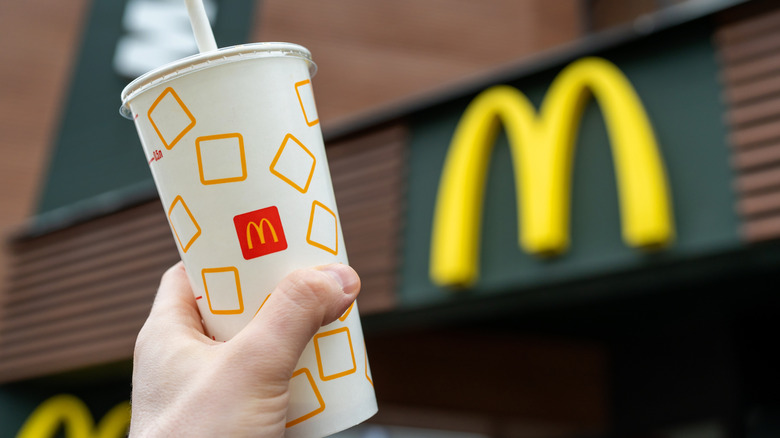 A hand holds up a McDonald's beverage cup with a McDonald's restaurant sign in the background