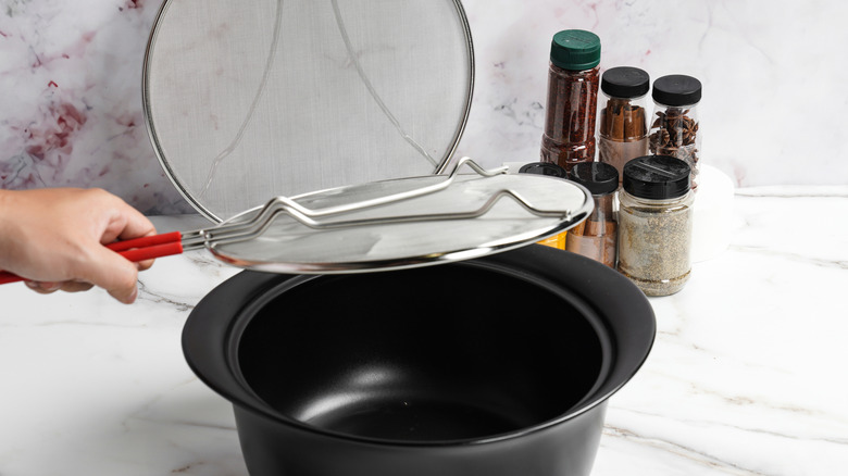 A person putting a splatter shield over a crock pot against a white marble countertop with spices in the background