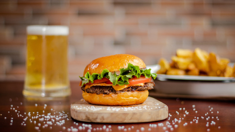 A smashburger sitting on a wooden serving board in front of french fries and a pint of beer