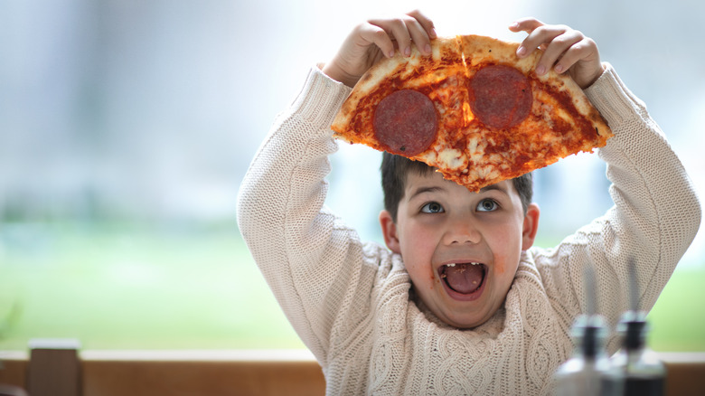 A young child with their front teeth missing joyfully holds up two slices of pepperoni pizza