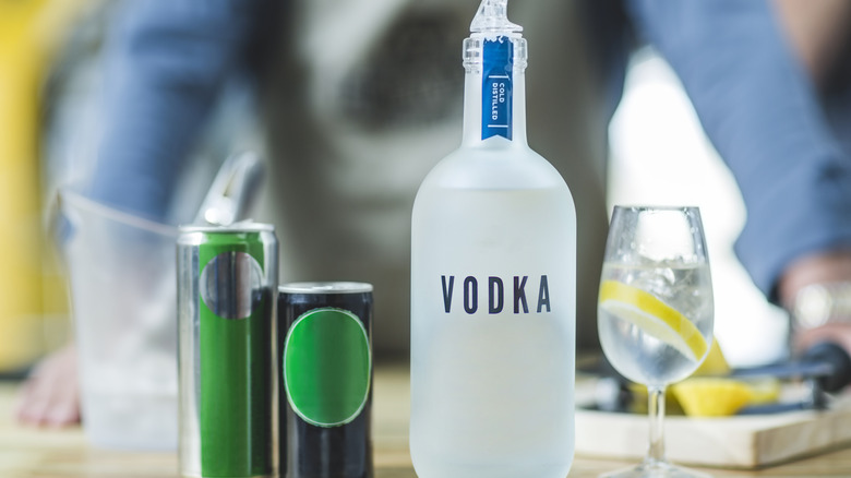 A bottle of vodka sits on a table next to several unmarked cans and a wine glass with ice and lemon wedge