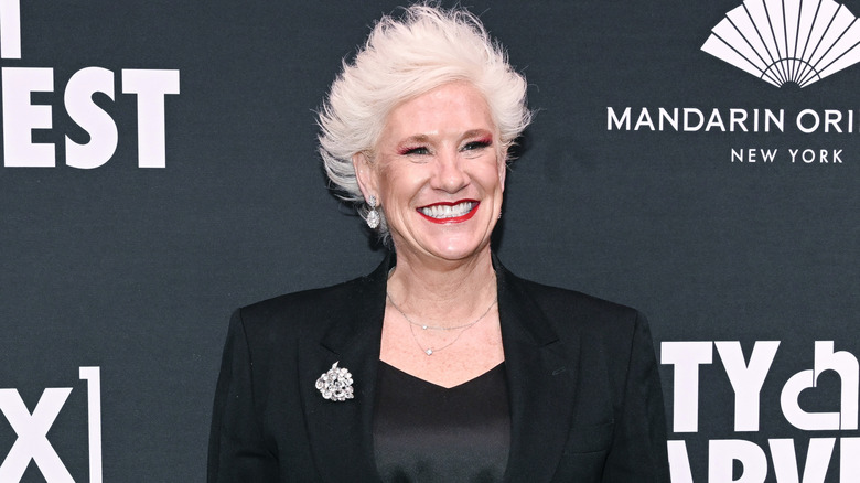 Anne Burrell smiles in a black blazer with a black top and red lipstick