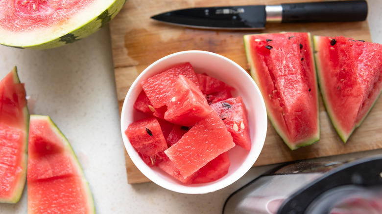 A watermelon has been thoroughly carved, with slices on a cutting board sitting next to a bowl of freshly trimmed watermelon