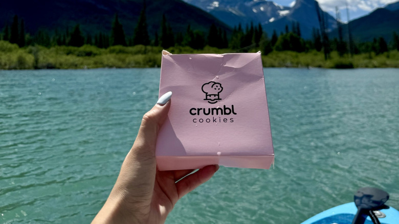 A kayaker holds a Crumbl Cookies box in front of the great outdoors, a river, some trees, and majestic mountains in the distancec