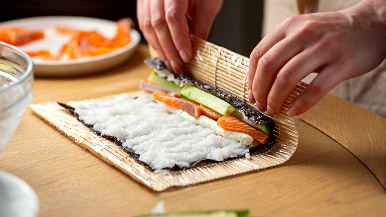 Someone rolls a homemade sushi roll featuring salmon, cucumber, and cream cheese
