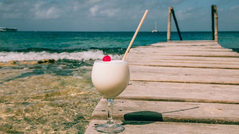 A pina colada sits on a jetty on a beautiful sunny beach next to clear blue waters