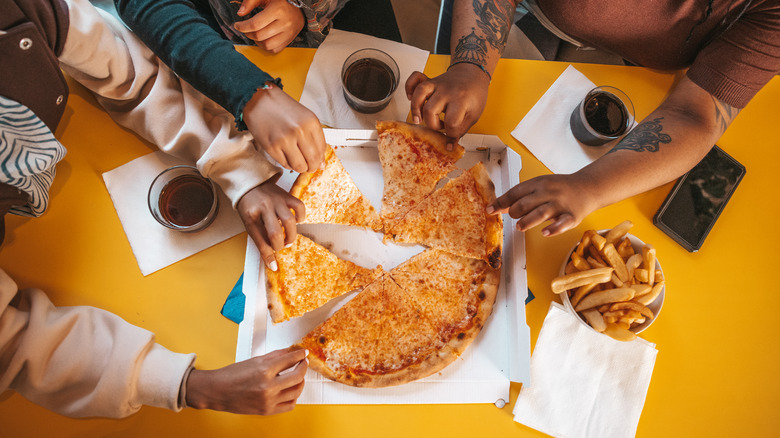 friends share a cheese pizza, fries, and colas at a food court table
