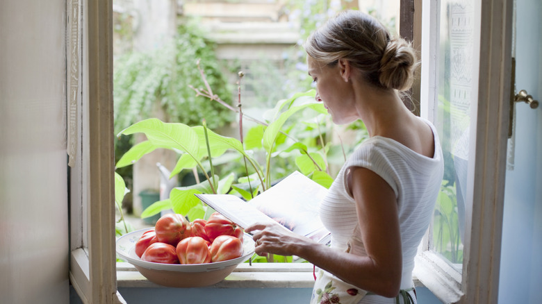 A woman looking out a window with a cookbook, with a bowl of tomatoes on the window sill
