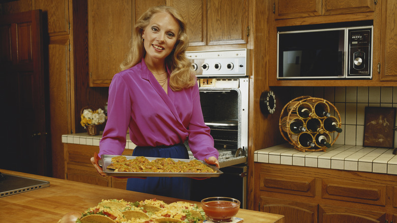 Vintage photo of a homeowner holding a tray of food in a circa 1980s kitchen featuring honey oak cabinetry, tiled countertops, and a retro microwave