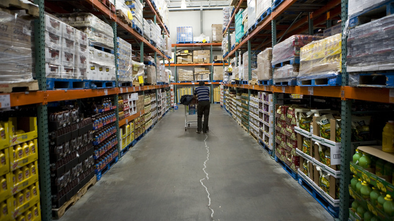 Someone shops at Costco, pushing a cart down a long aisle of canned and packaged foods