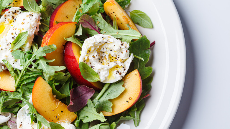Arugula salad with peach halves, burrata, basil, olive oil, and fresh cracked black pepper on a white dish