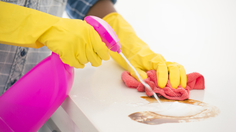 Hands in yellow cleaning gloves spray liquid from a pink bottle onto a countertop stain and wipe with a microfiber cloth