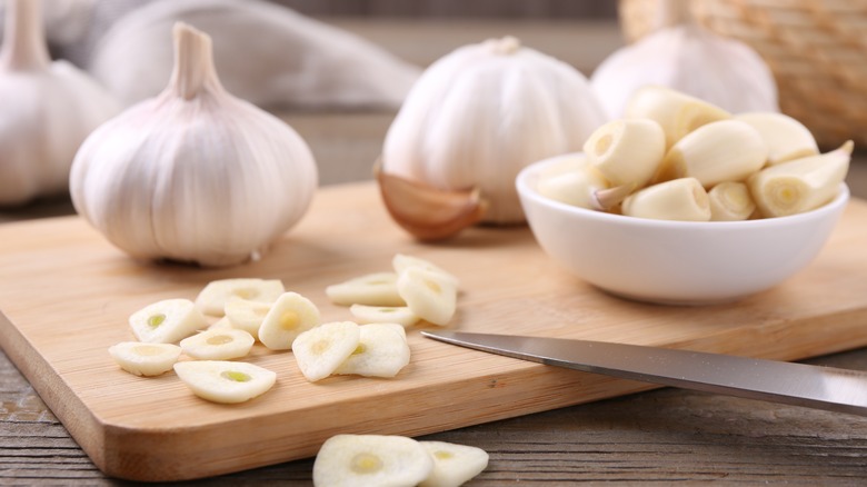 Sliced garlic on a wooden cutting board with bulbs of garlic, a small bowl with peeled cloves, and a knife blade around them