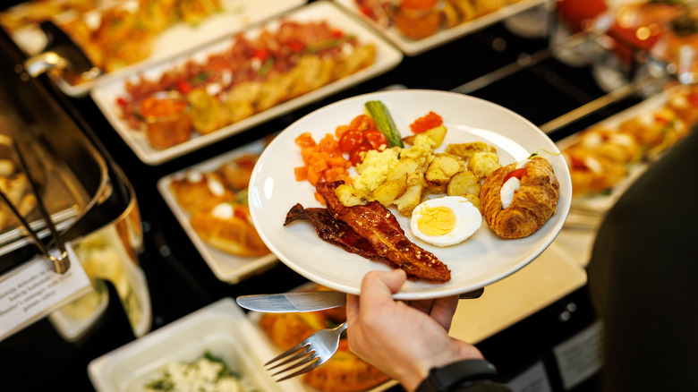 A hand holds a plate filled with breakfast foods above a buffet table