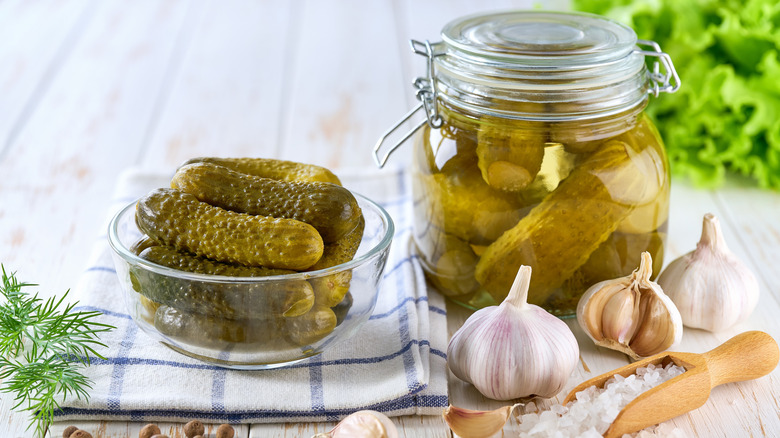 Jar and bowl of pickled cucumbers sit on a table next to salt, garlic, herbs, and peppercorns
