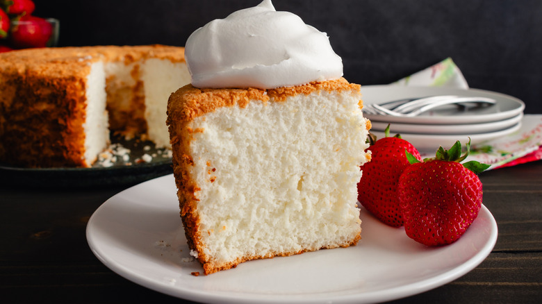 A slice of angel food cake topped with whipped cream sits on a white plate, with two whole strawberries sitting on the plate next to it