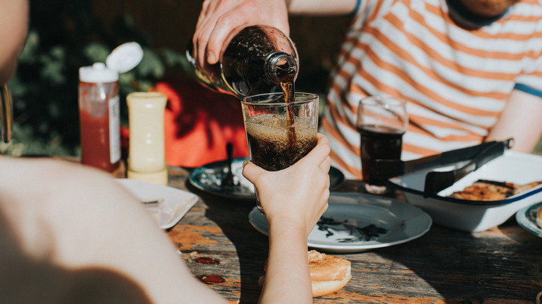 Someone pouring a glass of soda from a bottle at a sunny picnic
