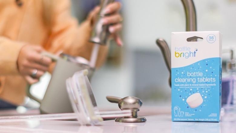 A box of Bottle Bright cleaning tablets on kitchen counter as someone uses sink in background
