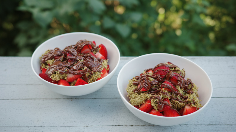 Two bowls sit on a picnic table outside, filled with sliced strawberries and topped with kataifi, pistachio cream, and drizzled chocolate