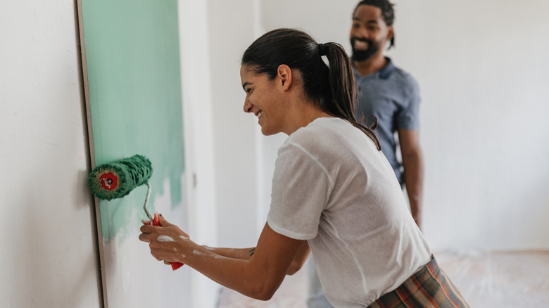 A smiling woman rolls a green paint color onto a wall while a man watches in the background