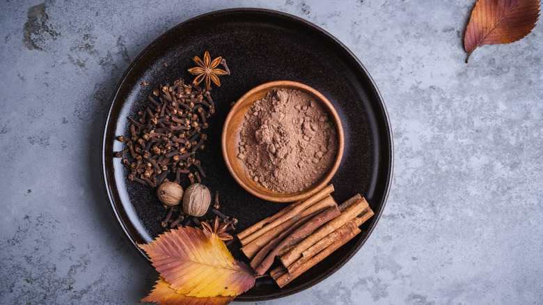 Various fall spices, including cinnamon sticks and star anise, sit on a black plate, with fall leaves adorning and in the background