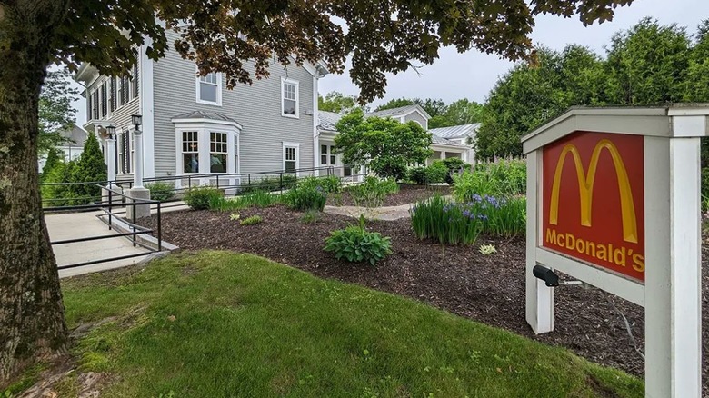 White wooden sign with red and gold McDonald's logo in gardened lawn area, with grey clapboard house in the background