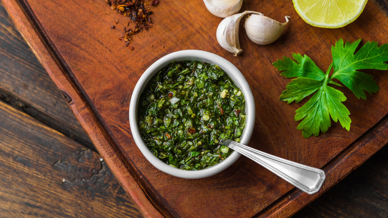 Chimichurri in a white dish with a spoon in it on a wooden cutting board and table surrounded by garlic, lime, pepper flakes, and an herb