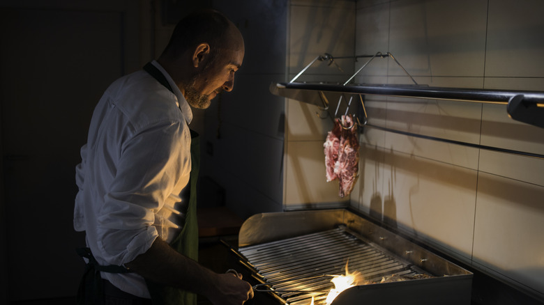 A chef tends to the kitchen grill while meat hangs over it