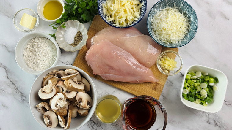 raw chicken on cutting board, two bowls of cheese, mushrooms, and other chicken Lombardy ingredients on a marble surface