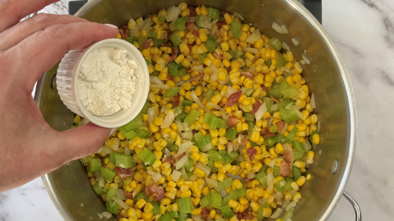 Hand holding small bowl of flour over soup pot with mixed veggies