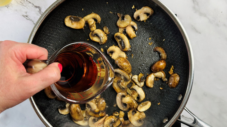 hand holding measuring cup of brown liquid over mushrooms in frying pan