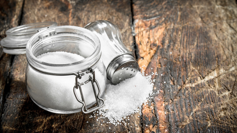Salt sits in a lidded jar and pours out of a shaker on a wooden surface