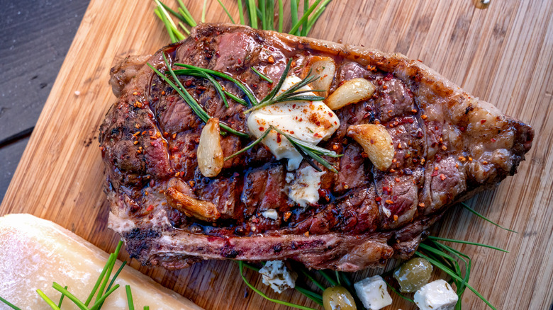 A piece of grilled bone-in ribeye steak rests on a wooden cutting board with rosemary, garlic, and butter