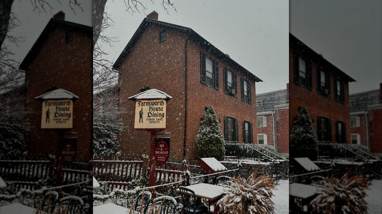 The Farnsworth House restaurant, a historic brick building, stands before a gray sky as snow gently falls and covers the ground