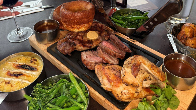 A feast laid out on a table consisting of steak, roast chicken, yorkshire puddings, jacket potatoes, mash, and steamed greens including broccolini