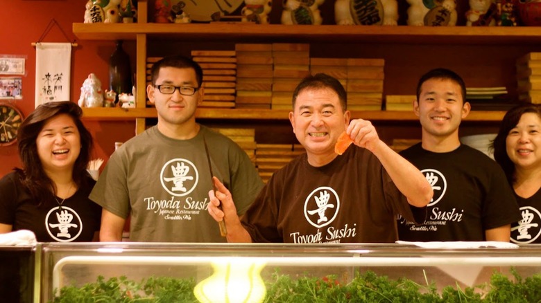 Natsuyoshi and his family, he is holding a piece of fish in one hand and a chopstick in the other, they are all smiling