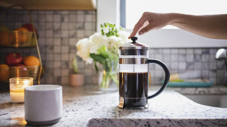 Someone pushes down the plunger on a coffee press that rests on a counter, with a pretty bouquet of flowers in the background
